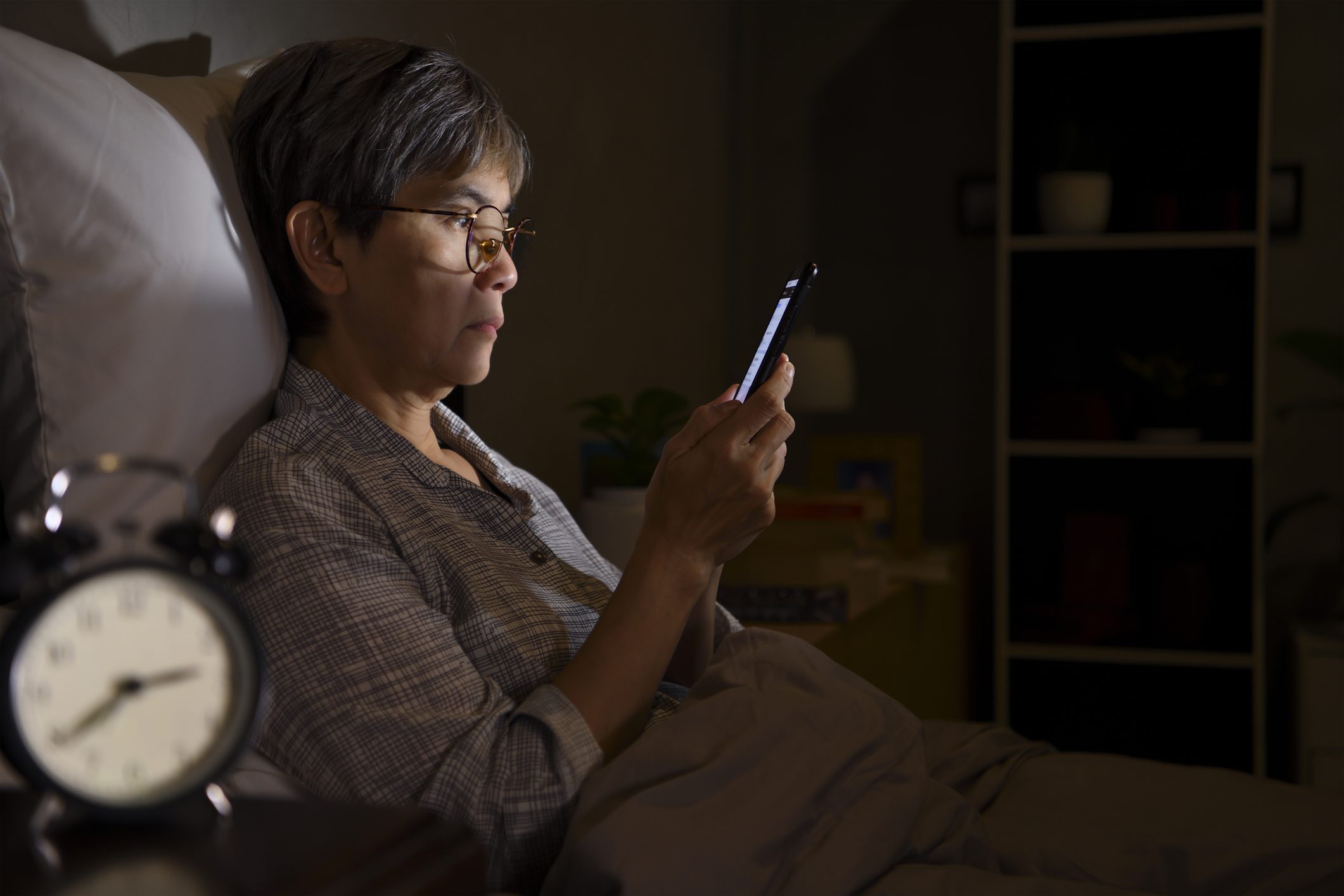 Asian senior woman having sore and tired eyes when using a smartphone while lying in bed at night